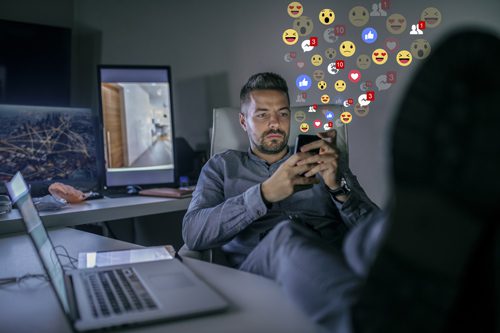 man at his desk with his feet kicked up, using his smart phone to access multiple social media accounts at a time - internet or social media addiction - substitute addictions