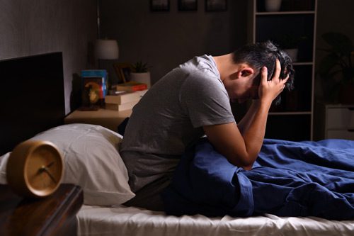 man sitting up in bed with his hands on the sides of his head - insomnia and addiction
